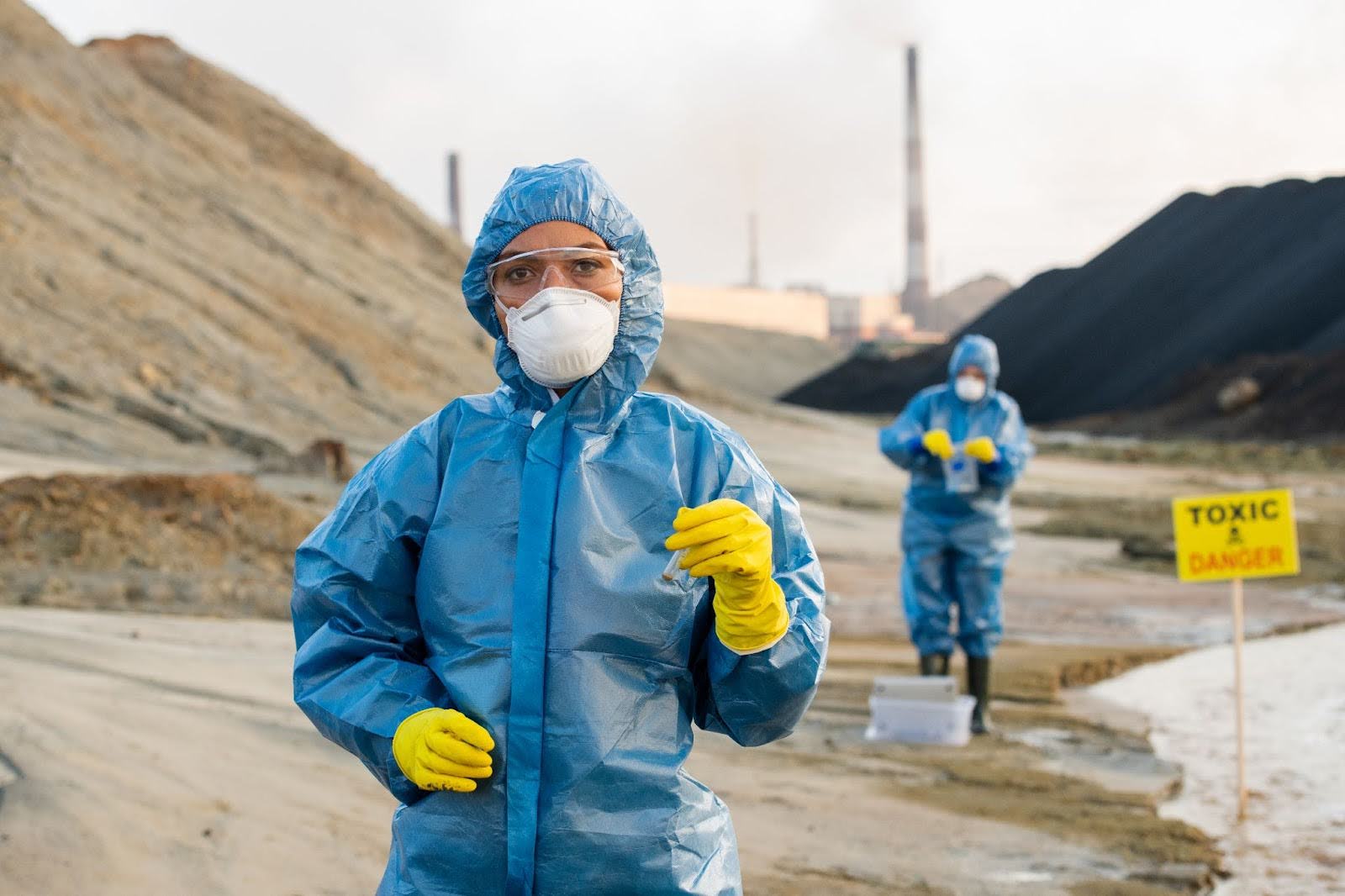 Two environmental scientists in blue hazmat suits work at a polluted industrial site