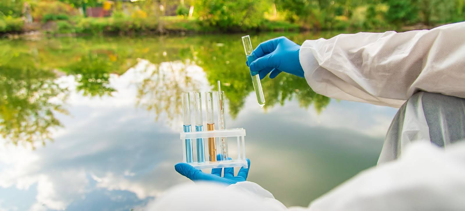 A scientist wearing a white protective suit and blue gloves examines a water sample in a test tube while holding a rack of other samples